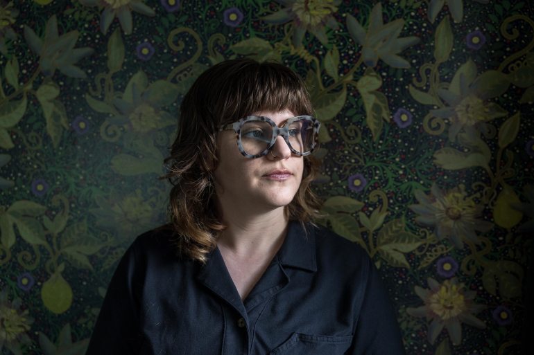 A portrait of a woman in front of a dark green floral wallpaper.