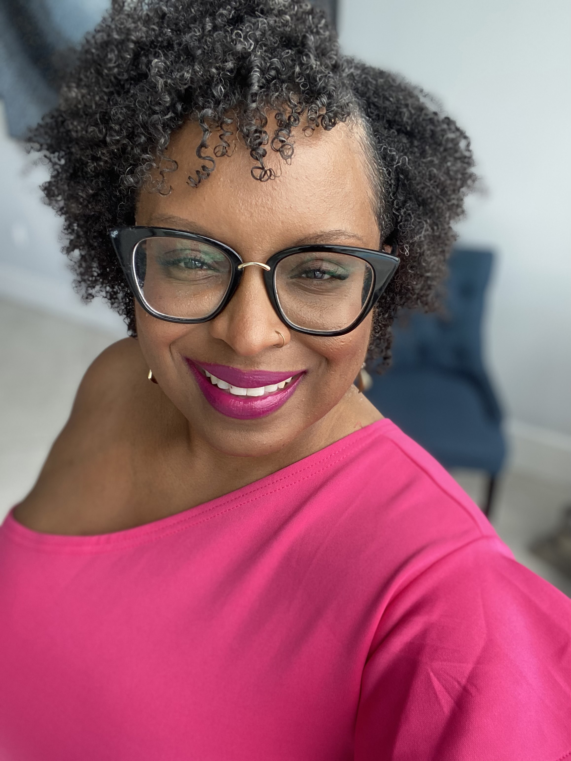 Cynthia Johnson is seen taking a selfie and smiling. A blue chair is seen blurred in the background behind her.